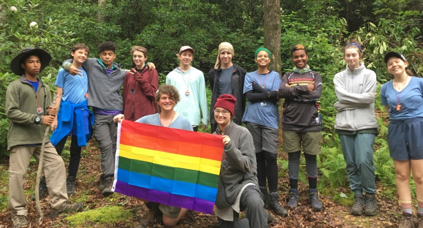 A group of young people standing in a wooded area hold a pride flag